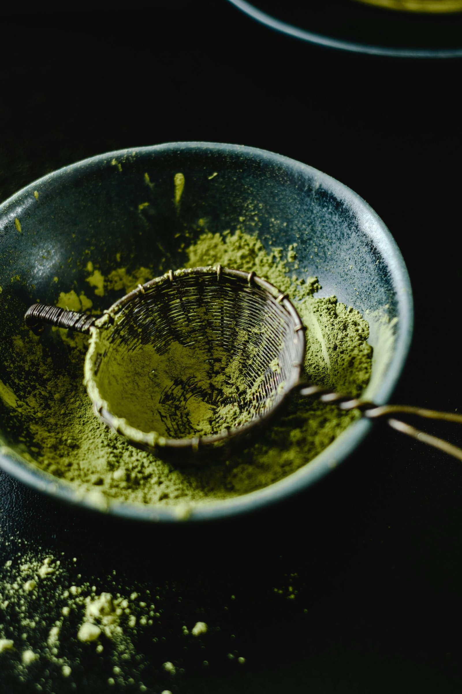 Close-up of matcha powder in a ceramic bowl with a bamboo sieve on dark background.